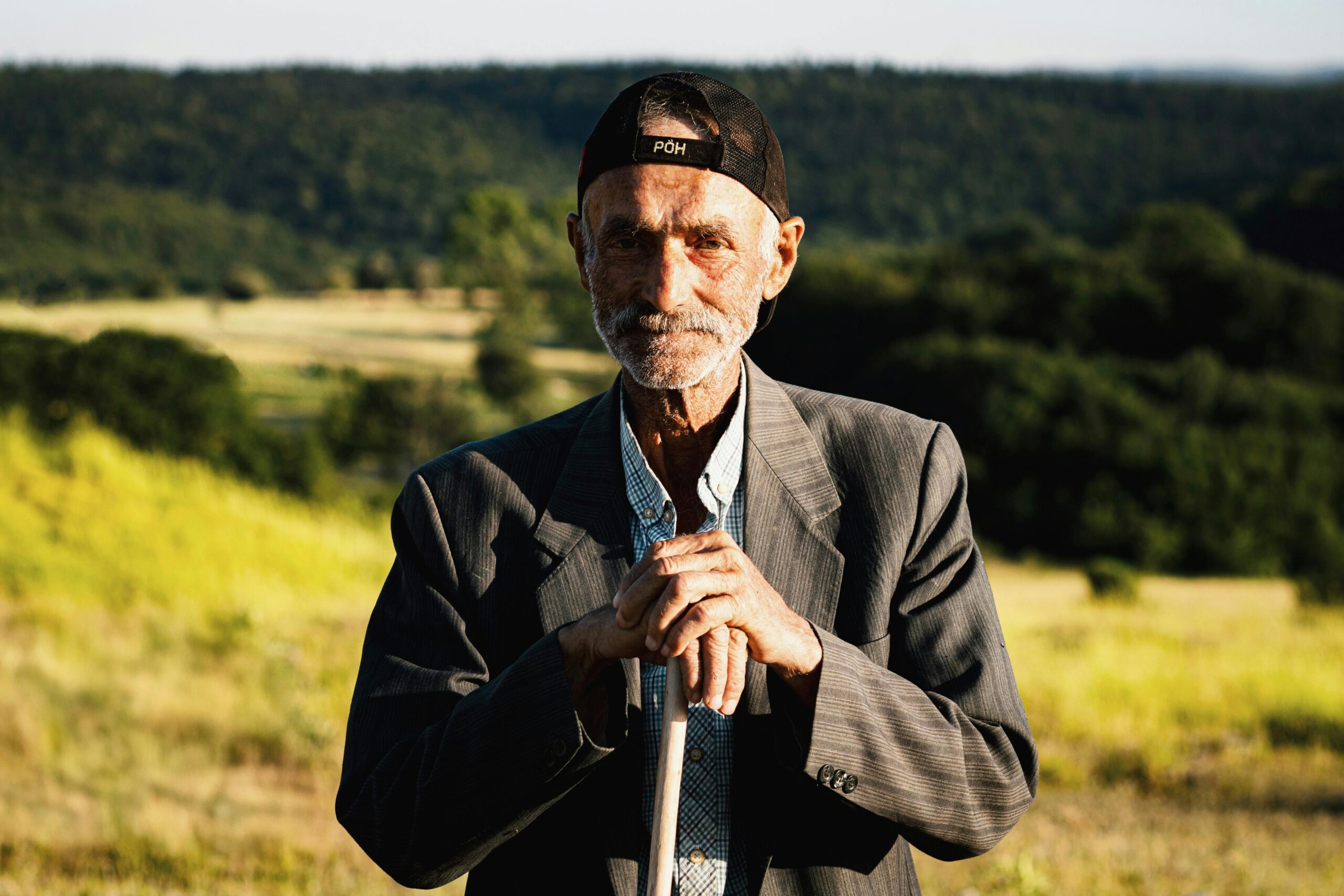 Elderly man in a suit and cap standing outdoors against a serene rural backdrop.